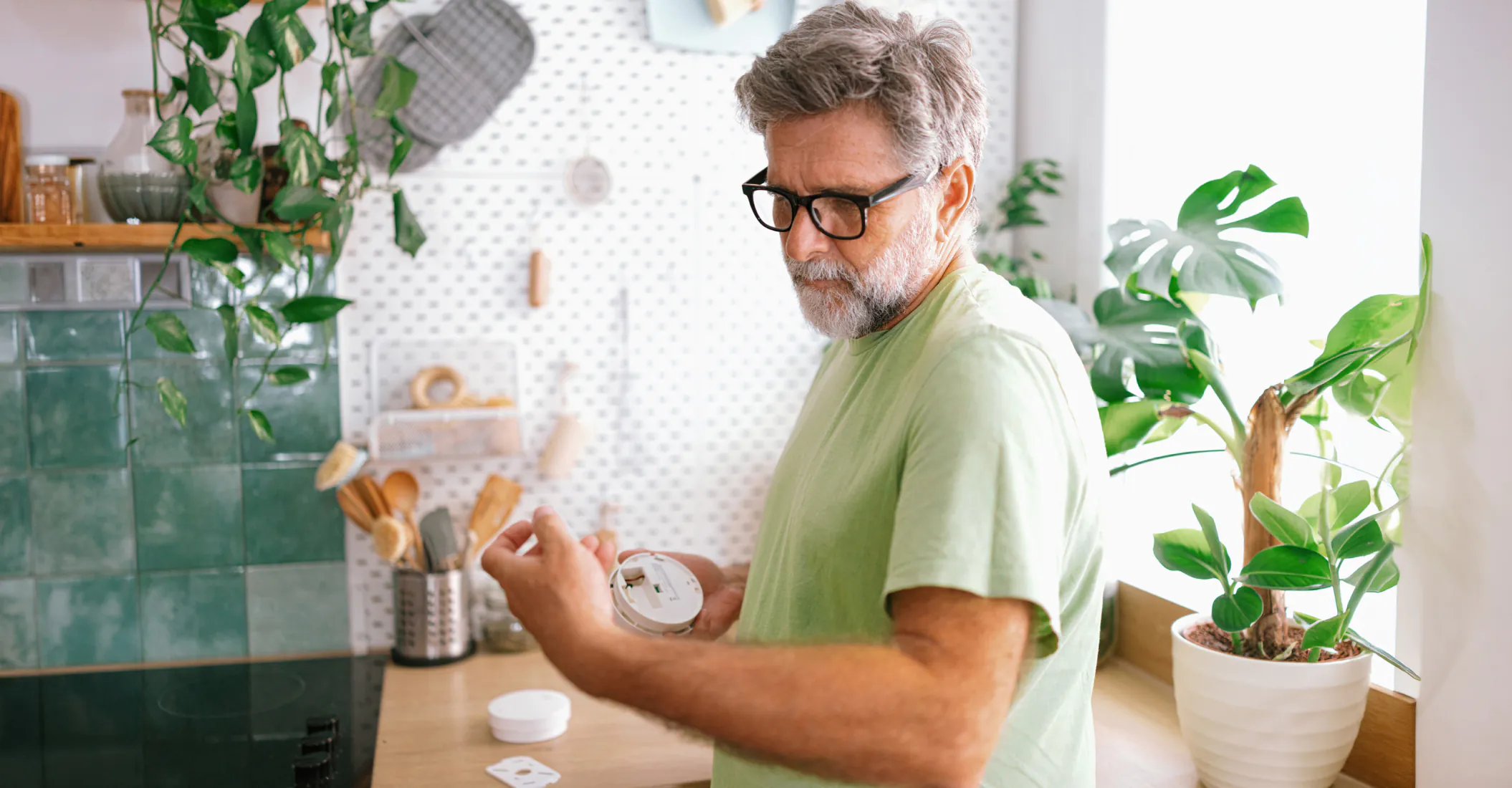 Older man wearing glasses holds a smoke detector and battery in a kitchen, appearing to inspect or change the battery. Green plants and kitchen utensils are visible in the background.
