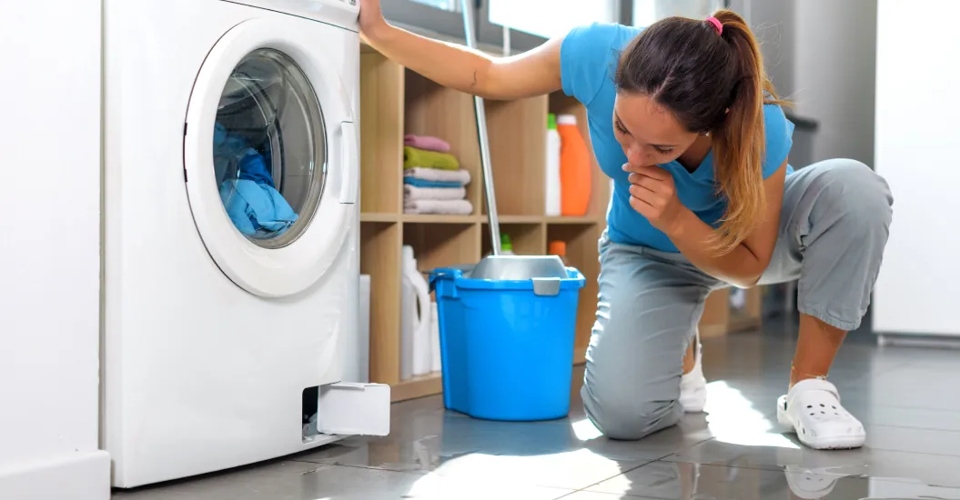 A woman inspects a leaking washing machine as water spills onto the floor; a mop and bucket are nearby