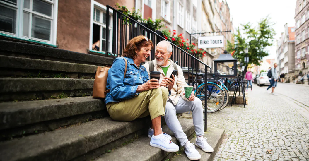 Two older adults sit on outdoor steps, smiling and holding coffee cups, with bicycles and a “Bike Tours” sign visible in the background.